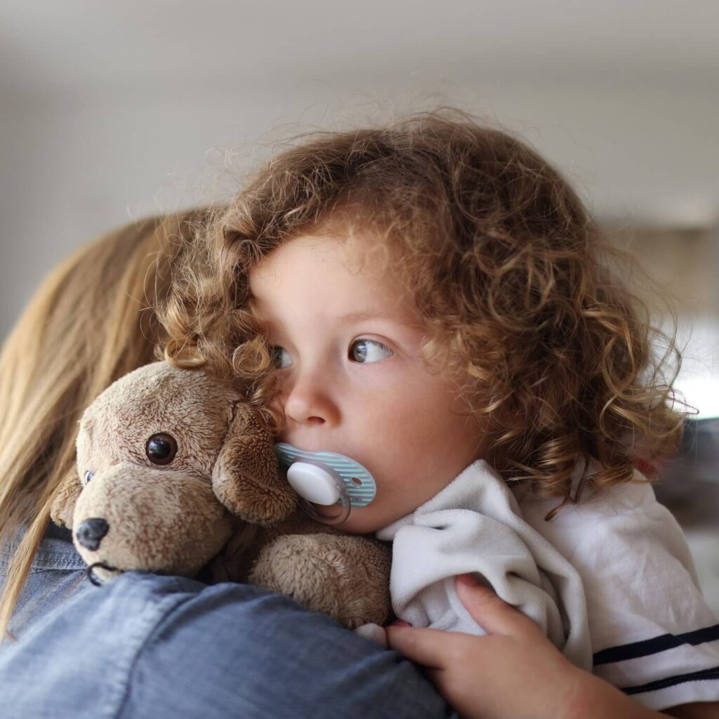 infant-with-pacifer-square An infant with a pacifer in her mouth is pictured here holding a stuffed animal while on her mother's shoulder.