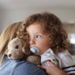 An infant with a pacifer in her mouth is pictured here holding a stuffed animal while on her mother's shoulder.