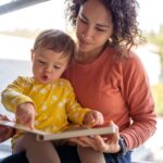 A mom is holding her infant child as the little one uses her index finger to point to a page in a book.